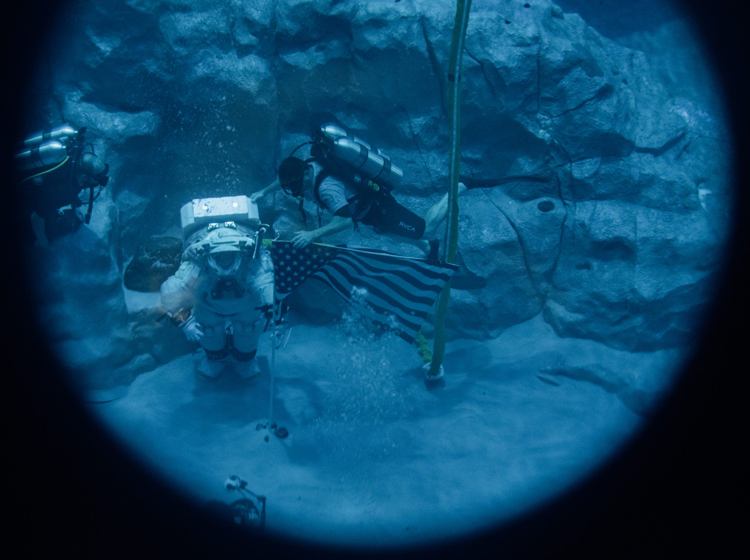 Un astronauta che indossa una tuta spaziale cammina sul fondo di una piscina accanto a un sub che indossa una maschera da sub