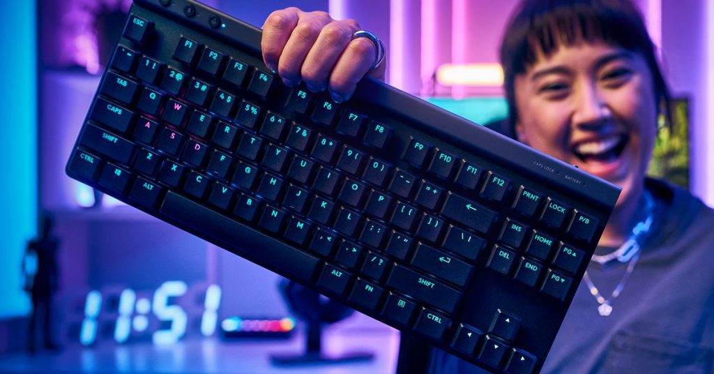 A user holds the Logitech G515 keyboard in one hand in front a computer setup featuring neon lighting.